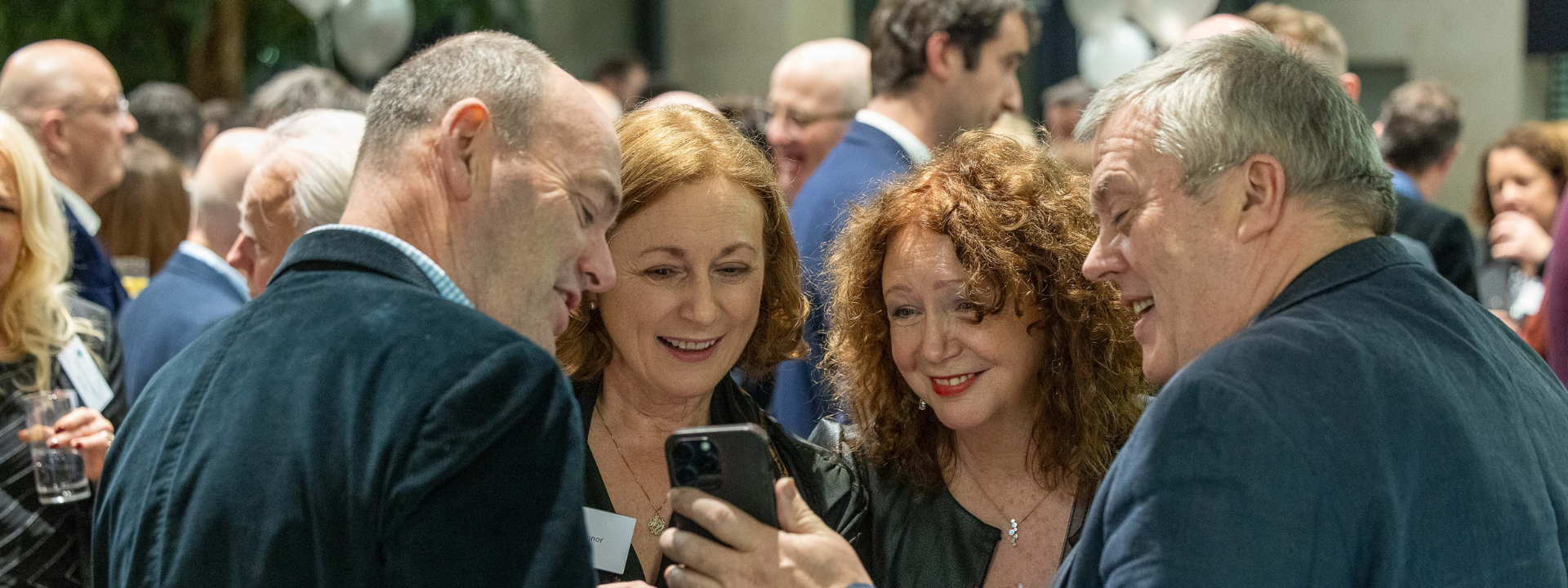 Four alumni at their reunion, looking at a phone screen and smiling.